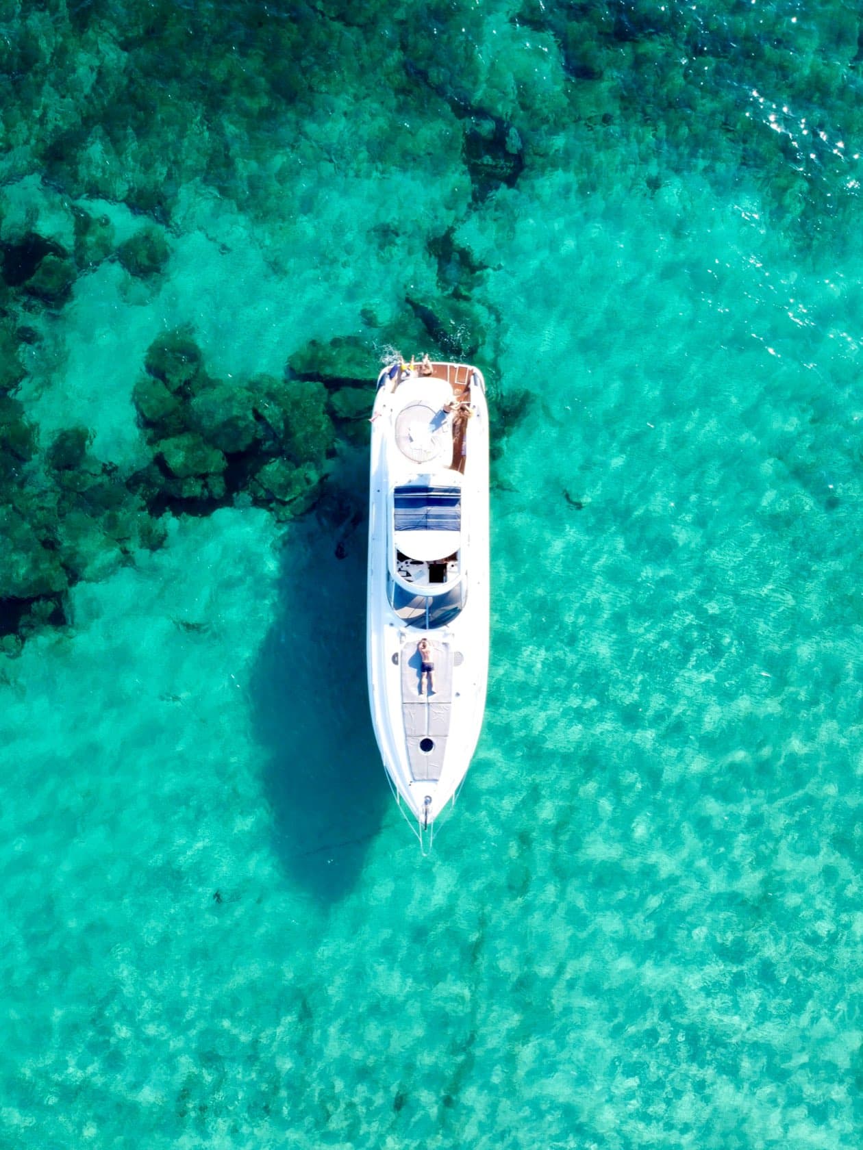 Yate de motor fondeado en agua turquesa, visto desde arriba, con alguien en el asoleadero de proa